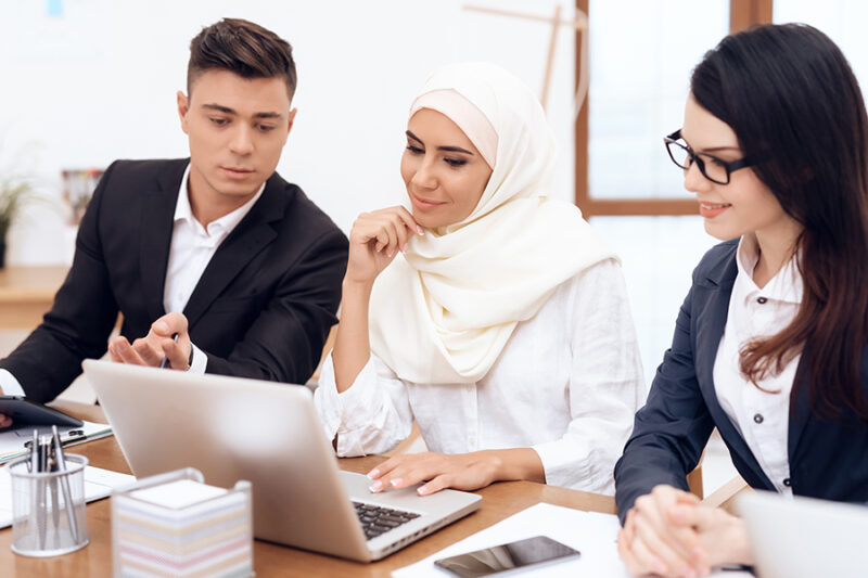 The Arab woman in hijab works in the office together with her colleagues. They are engaged in business. On the desk is a laptop.